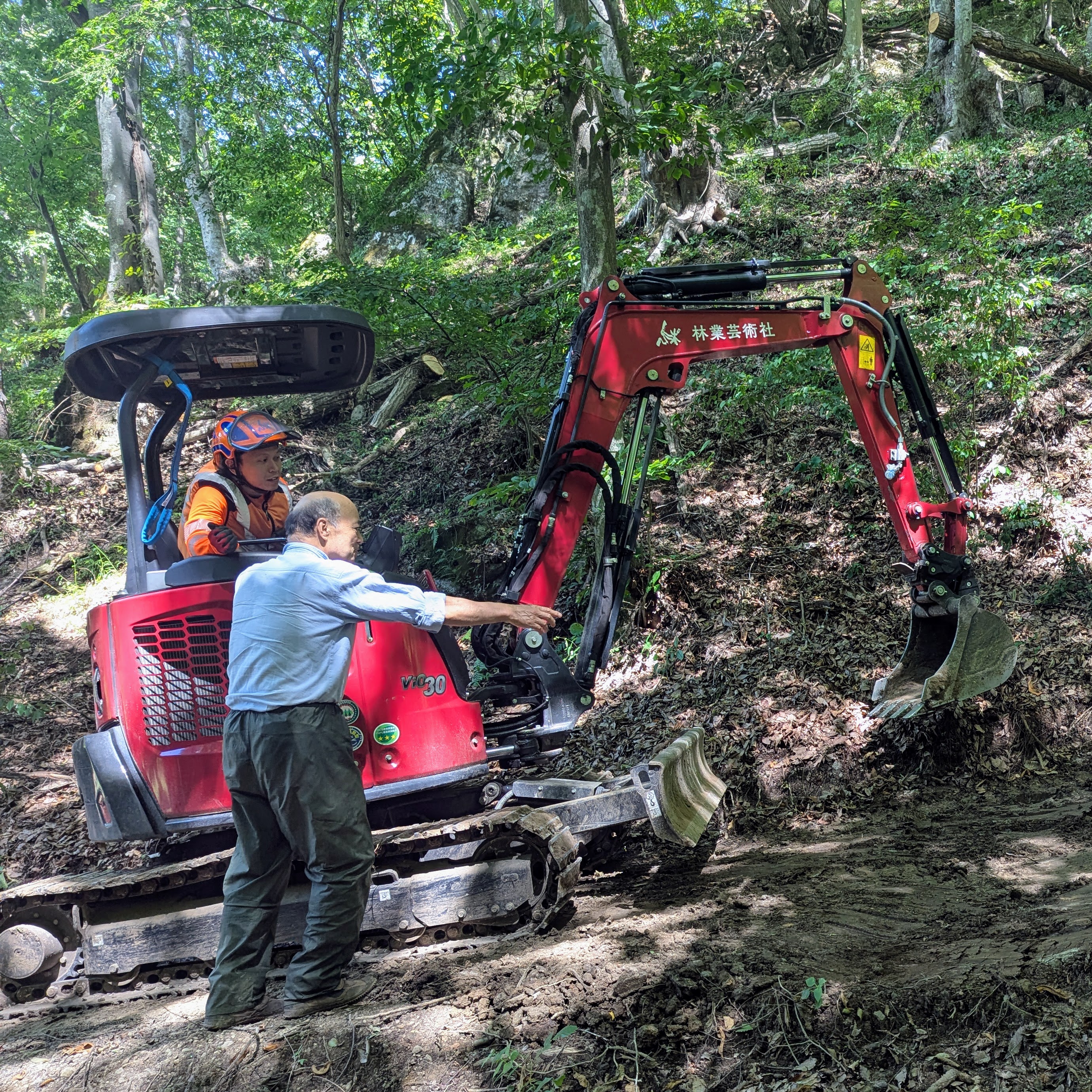 林業芸術社フィールドの山道で、バックホウを操作しながら指導を受ける参加者の様子